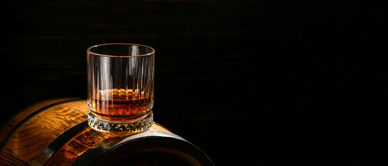 Wall Mural - Glass of whiskey on wooden barrel against dark background with space for text