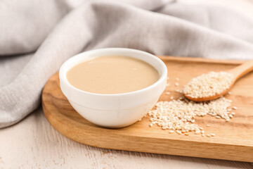 Board with bowl of tasty tahini and sesame seeds on light wooden table, closeup