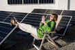 © rh2010 - Young woman talks on phone while sitting relaxed on rooftop with a solar station. Happy owner of energy-independent household. Eco-friendly and sustainable lifestyle