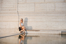 Woman Sitting By Pond Against Wall In City During Sunset
