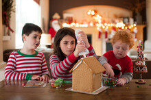 Three Kids Decorating A Gingerbread House At Home For The Holidays