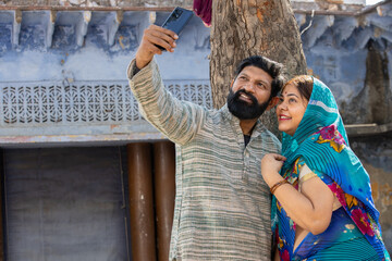 Wall Mural - Young happy rural indian couple wearing traditional outfit sitting on bed taking selfie picture with smartphone .