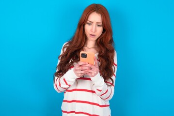 Wall Mural - young woman wearing striped sweater over blue background looks with bugged eyes, holds modern smart phone, receives unexpected message from friend, reads reminder.