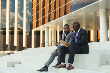 Sticker - Friendly meeting of business partners outdoors. Two dark-skinned men in suits sit on the steps of a city building with a notebook and have a conversation. Working break. Support and collaboration