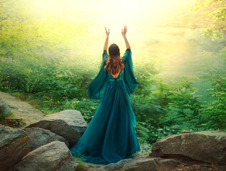 Fantasy woman queen prays in summer forest hands raised to divine magical sun light sky. Girl in long silk green dress fabric flying in wind. Mystery lady goddess back rear view enjoying life harmony 
