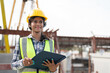 © Supachai - Asian woman engineer holding document smiling at construction site. Confident female Indian wearing protective helmet and vest working in factory making precast concrete wall for real estate housing.