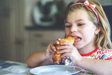 Wall Mural - Smiling child at breakfast. Food and happy kids. The girl is eating a croissant. Cute preschool girl having healthy meal.