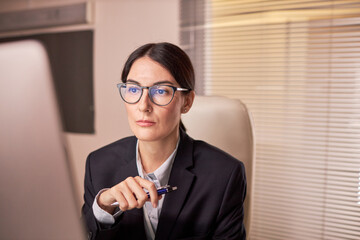 Wall Mural - Portrait of serious businesswoman wearing glasses and looking at computer screen while working at night in office, copy space