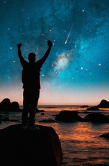 Wall Mural - silhouette of a person on a rock hands up at night with milky way and stars on the background over the ocean