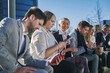 © Oscar - young coworkers during break time outside of office, sitting on bench having fun using modern smartphone, dressed in fashionable elegant clothes. selective focus of asian woman looking at camera