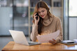 © David - Attractive successful young business Asian woman in striped blouse working in modern office, making phone call to potential client, having nice conversation, sitting at desk in front of open laptop