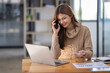 © David - Attractive successful young business Asian woman in striped blouse working in modern office, making phone call to potential client, having nice conversation, sitting at desk in front of open laptop