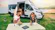 Happy young couple sitting outdoors in summer with camper van and beach on background