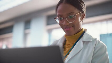 Poster - Portrait of Young Black Female Specialist in a Lab Coat Smiling and Using Laptop Computer. Professional and Successful Woman Working as an Engineer, Developing High Technology Projects