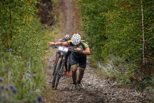Two Cyclists Go Uphill With Their Mountain Bike. Heavy Climb On Muddy Slope. Cross-country Cycling Competition