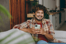 Young Happy Indian Man Wears Casual Clothes Hold In Hand Use Mobile Cell Phone Sits On Grey Sofa Couch Stay At Home Hotel Flat Rest Relax Spend Free Spare Time In Living Room Indoor. Lounge Concept.