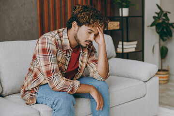 Wall Mural - Side profile view young sad upset displeased Indian man wears casual clothes prop up head sits on grey sofa couch stay at home hotel flat spend free spare time in living room indoor. Lounge concept.