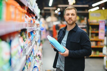 Young man in the supermarket in the household chemicals department. Large selection of products. A brunette in a glasses and a beard in a beige coat.