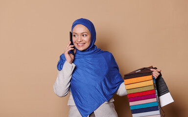 Charming Arab Muslim woman with head covered in blue hijab, confident successful sales manager, decorator talks on mobile phone, taking orders for sofa upholstery materials, isolated beige background