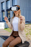 Fototapeta Sypialnia - Serious businesswoman testing virtual reality while sitting alone on street in front of office building. Beautiful female entrepreneur with VR headset making swipe and expand gestures.
