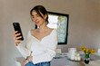 © PhotoBook - Lovely cute girl with dark collected hair wearing white shirt is using smartphone with happy smile and standing in the living room in sunny warm day.