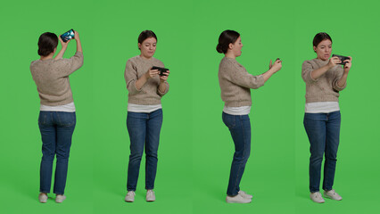 Poster - Female model playing video games online on smartphone, standing over greenscreen backdrop and having fun. Young adult enjoying shooter gaming competition play in studio, positive woman.