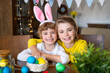 © Chiralli - Easter Family traditions. Two caucasian happy children with bunny ears dye and decorate eggs with paints for holidays while sitting together at home table. Kids embrace having fun together