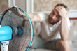 © Евгений Шемякин - Young bearded man using electric fan at home, sitting on couch cooling off during hot weather, suffering from heat, high temperature