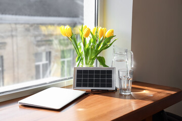 Wall Mural - Portable solar panel charging laptop, jug of water and vase with tulips on table near window