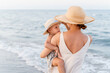 © Cavan Images - Woman in a straw hat holding a little girl in her arms near the sea