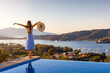 © moofushi - A happy holiday woman in a dress stands at the swimming pool and enjoys the summer sunset behind the mediterranean sea