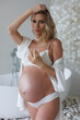 Beautiful pregnant woman in white retro underwear and gown standing near bath. Young blond lady sitting on sofa in spacious room with big windows. Big pregnant naked belly. Pretty woman waiting child.