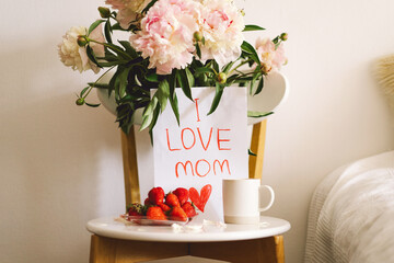 Breakfast for Mothers Day. Heart shaped white plate with fresh strawberries, cup of coffee, gift and Peonys bouquet with gift in bed. Still life composition. Happy Mother's Day.