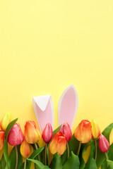 Wall Mural - Easter concept. Top view vertical photo of rabbit bunny ears in tulips flowers and colorful easter eggs on isolated yellow background with empty space