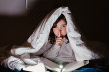 Cute Girl Reading Book Under Blanket And Showing Secret Gesture