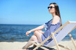 © rogerphoto - Happy young brunette woman relaxing on a wooden deck chair at the ocean beach while smiling and wearing fashion sunglasses. The enjoying vacation concept.