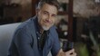 © nyul - Confident older white man at home, sitting on couch using phone in dark living room. Portrait of happy, mature age, middle age, mid adult caucasian man in 50s, smiling.