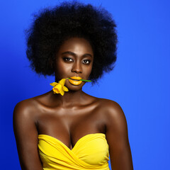 Wall Mural - Woman Beauty with Afro Hairstyle and Yellow Lipstick Make up over Blue Studio background. Dark Skin African Girl holding Spring Flower in Mouth. Women Makeup and Cosmetology
