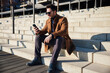 © baranq - Happy stylish man wearing coat sitting on stairs in the city using phone