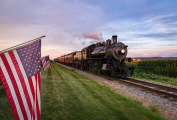 Canvas Print - A View of a Classic Steam Passenger Train Approaching, With American Flags Attached to a Fence on a Sunny Summer Day