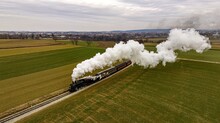 An Aerial View Of A Steam Passenger Train Approaching, Traveling Thru Open Farmlands, Blowing Lots Of White Smoke, On A Winter Day