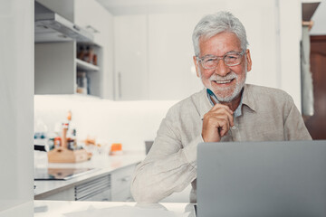 Aged remote worker. Concentrated senior male in glasses work on laptop from home office looking at the camera. Old age man employee freelancer sit at kitchen table by pc typing report online
