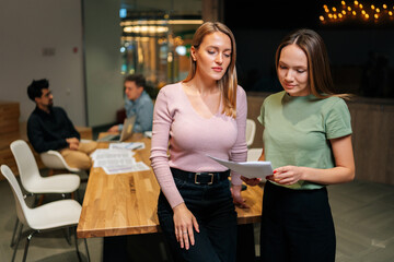 Wall Mural - Portrait of two pretty young businesswomen working, talking together, reading paper document, having informal meeting on background of startup business team chatting at desk on background.