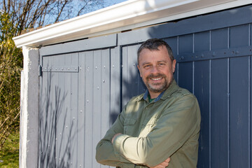 Outdoor portrait of happy handsome white man arms crossed in grey door wooden hut background in home garden