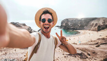 Handsome Man Wearing Hat And Sunglasses Taking Selfie Picture On Summer Vacation Day - Happy Hiker With Backpack Smiling At Camera Outside - Tourist Walking On The Beach - Traveling And Technology