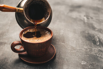 Wall Mural - Pouring black coffee drink from turkish jezve coffee pot into clay cup on dark background