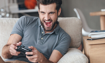 Poster - Excited young handsome man playing video game a while sitting on the couch at home