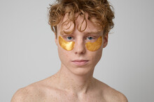 Close Up Of Curly Ginger Shirtless Male Model With Freckles And Blue Eyes Wearing Golden Gel Under Eye Patches And Looking At Camera. Male Beauty Routine, Male Skincare, Under Eye Bag Treatment.