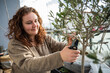 Woman with brown curly hair smiling while cutting the branch of a olive tree during spring with a secateurs on her balcony