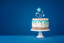 White Birthday Drip Cake With Teal Ganache, Star Toppers And Fun Candles Over Dark Blue Background